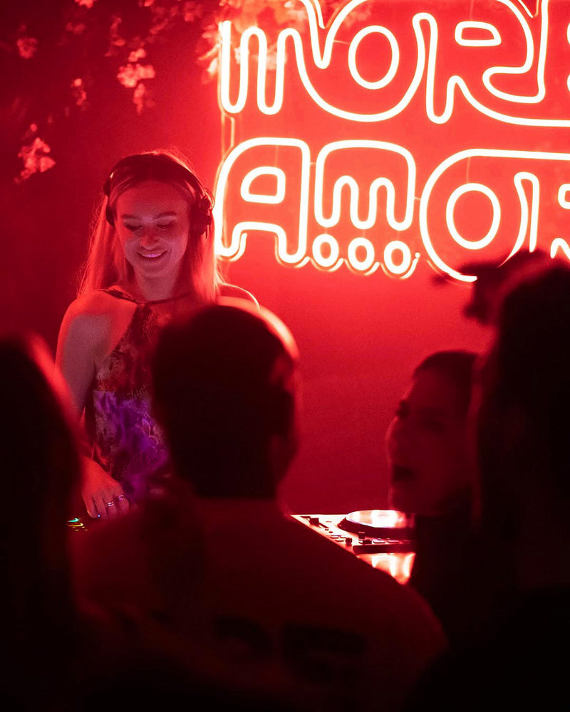 DJ at a party with neon sign 'More Amor' in the background